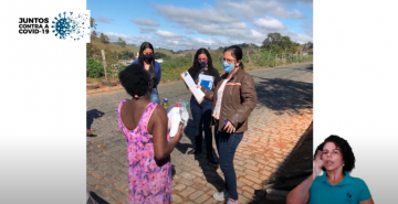 Três mulheres, de máscara, entregam um kit de higiene para uma mulher negra, de costas